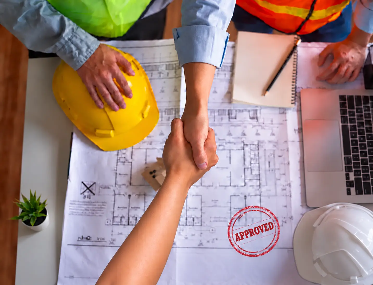 Handshake over construction plans stamped approved, with hard hats and a laptop on the table